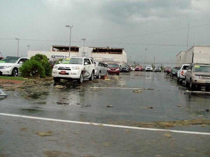Car lot with minimal damage, building window blown out in the background.